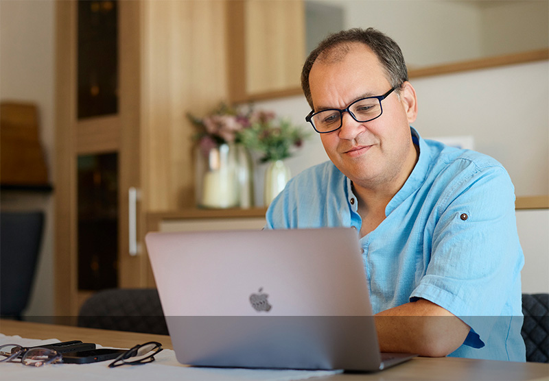 Ein Mann mit Brille sitzt am Küchentisch und arbeitet an seinem Macbook.