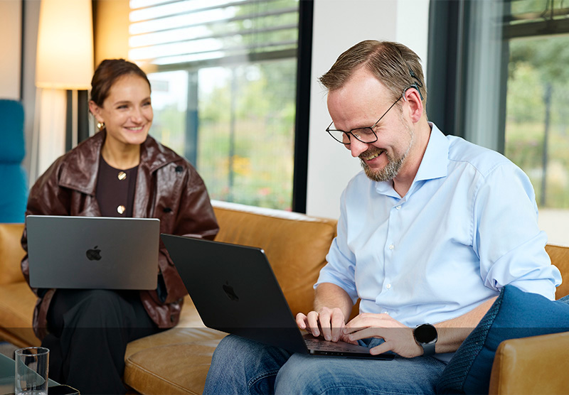 André Klein sitzt auf der Couch an einem Macbook mit einer Kollegin, die ebenfalls ein Macbook hat. Beide sind am Lachen.